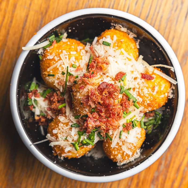 Photo of our Mac & Cheese Balls - These golden spheres of delight are enriched with Parmesan, chives, and bacon bits, making them a heavenly treat. If you’re looking for a larger portion, try our Mega Mac with the same mouthwatering flavors. Both options come with a luscious Chipotle Ketchup dipping sauce.