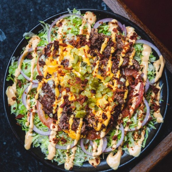 Our cheese Burger Salad features two smashed up smash patties, topped with American and Swiss cheese, shredded lettuce, ripe tomato, crunchy red onion, gherkin, and savory bacon bits.